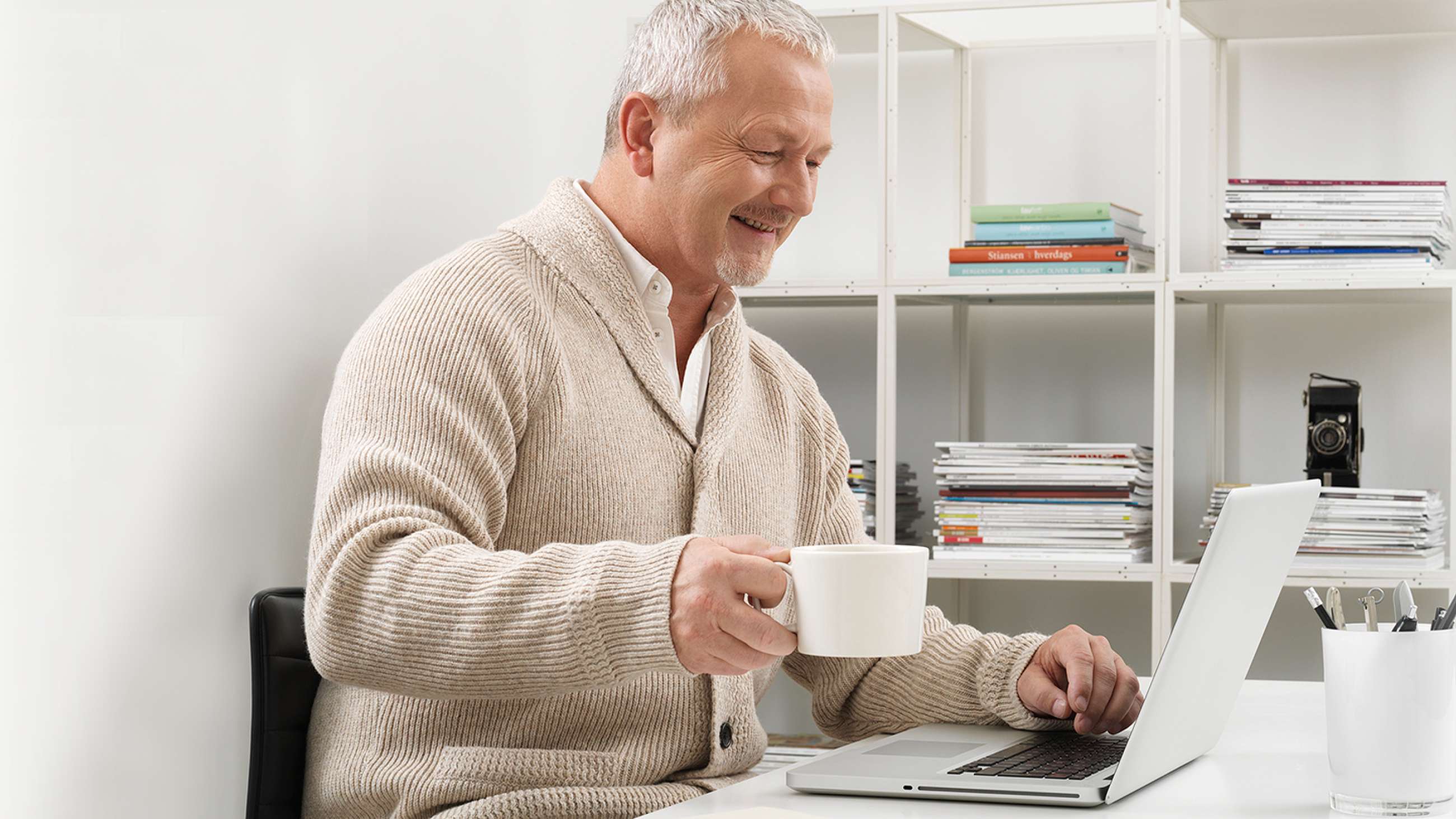 Mann holder en stor hvit kopp samtidig som han sitter foran sin laptop.