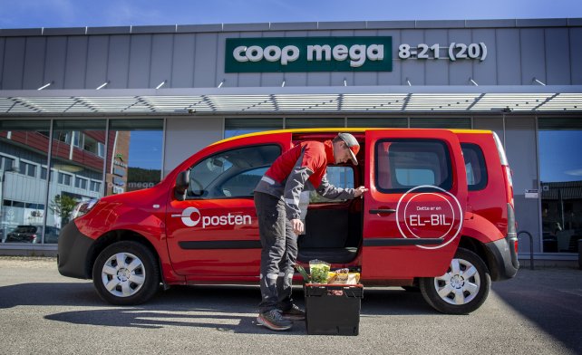 Postmann og elektrisk postbil parkert foran en Coop Mega. Foran seg har han en kasse med dagligvarer.
