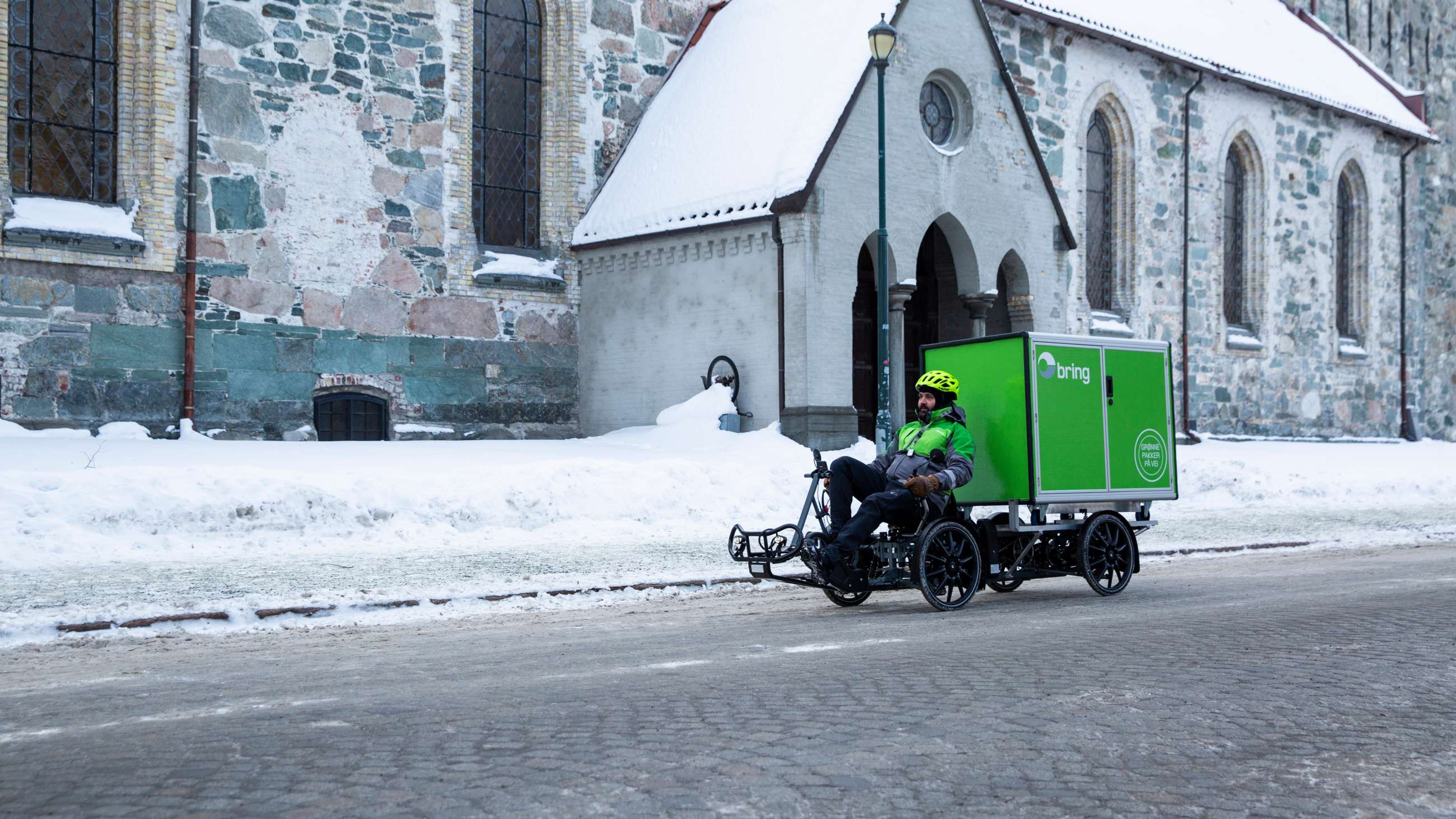 Mann i Bring-uniform sykler varesykkel med Bring-container bakpå. Vinterbilde fra Trondheim.