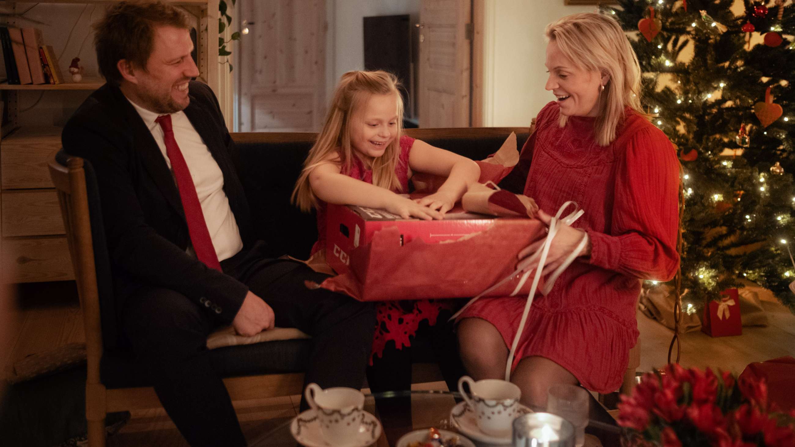 Et barn sitter mellom en mann og en kvinne i en sofa på julaften og åpner en julegave