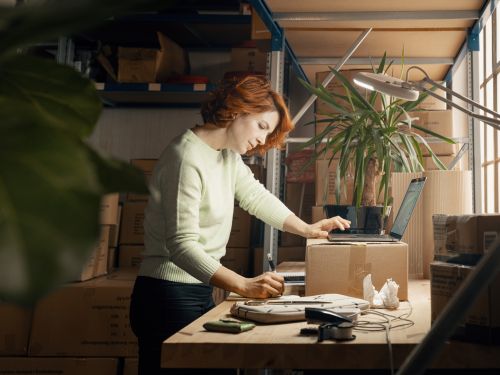 Kvinnelig nettbutikkinnehaver står ved en pappeske på et arbeidsbord med mange esker i bakgrunnen. Hun noterer på ei blokk mens hun holder den andre hånda på en oppslått laptop.