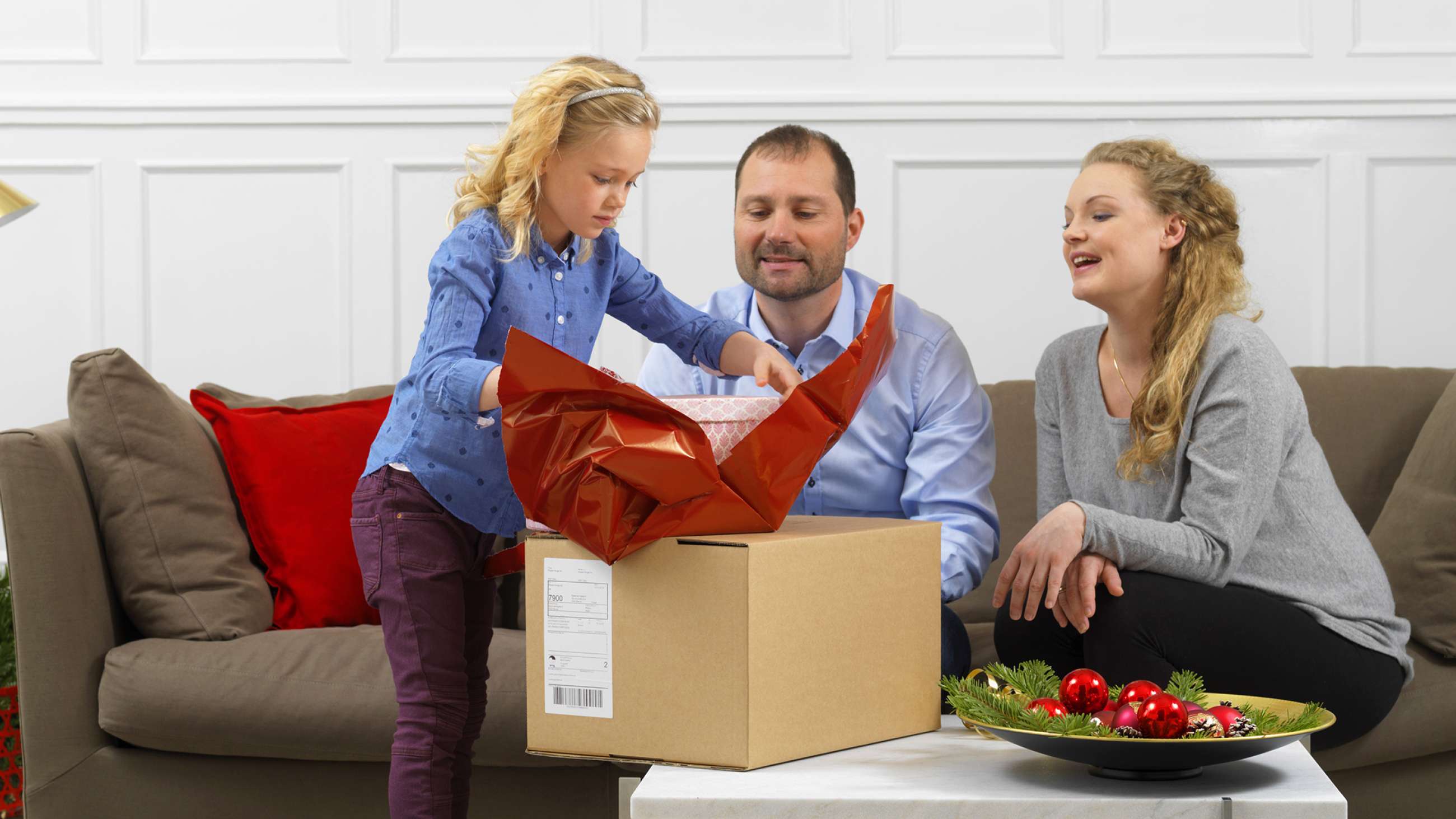liten jente åpner pakke foran sine foreldre som sitter i en brun sofa.
