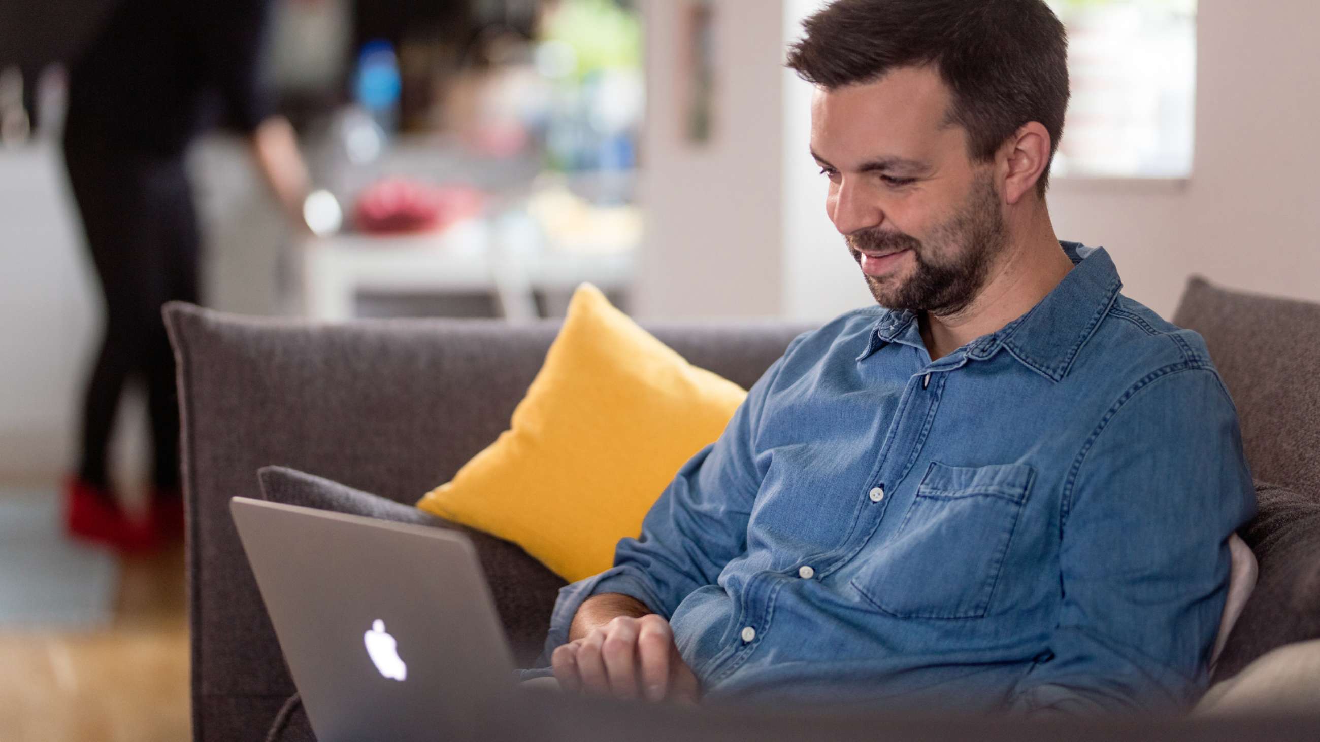 Ung mann med skjegg og jeansskjorte sitter smilende i sofaen, med en laptop på fanget