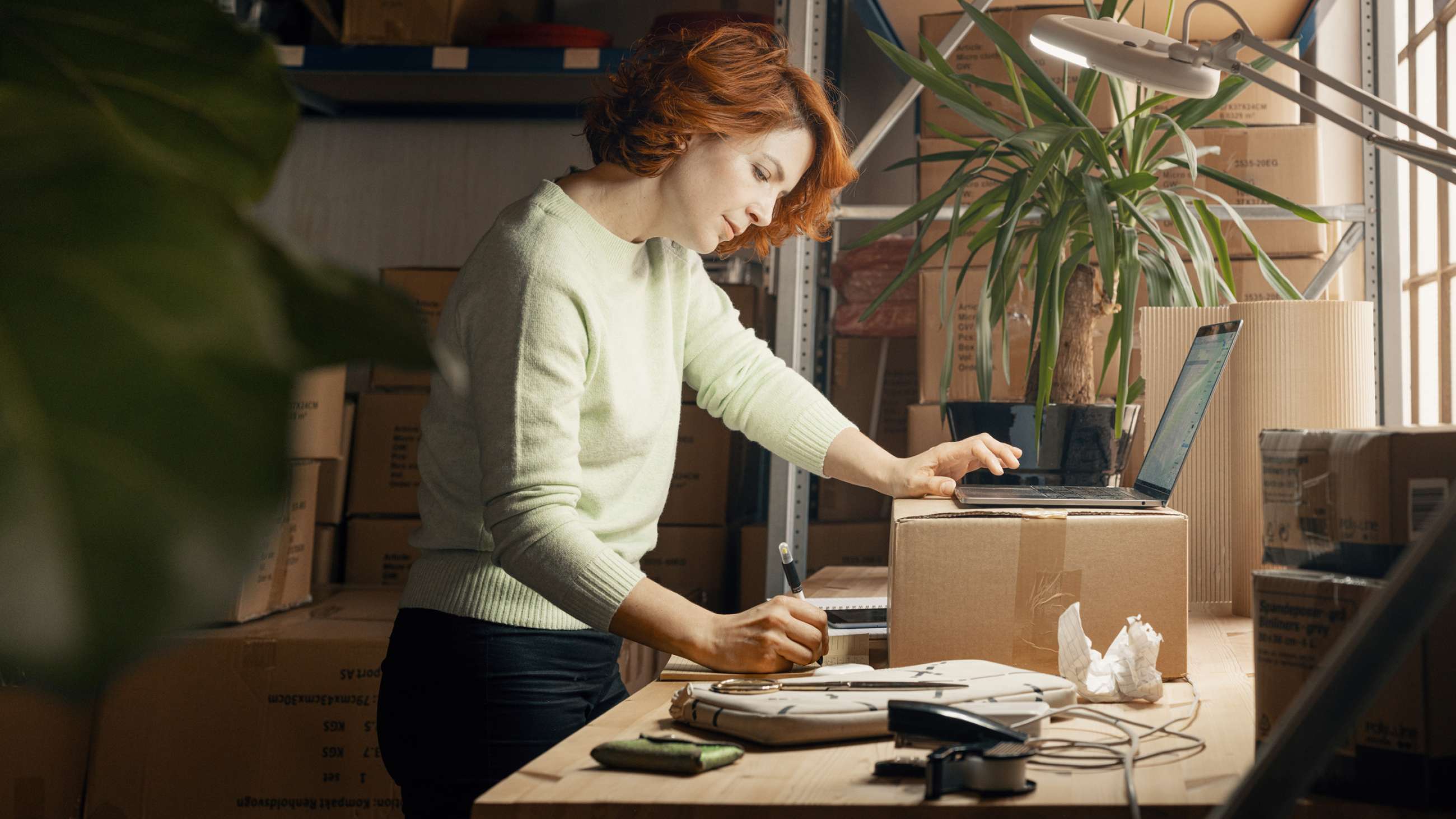Kvinnelig nettbutikkinnehaver står ved en pappeske på et arbeidsbord med mange esker i bakgrunnen. Hun noterer på ei blokk mens hun holder den andre hånda på en oppslått laptop. 