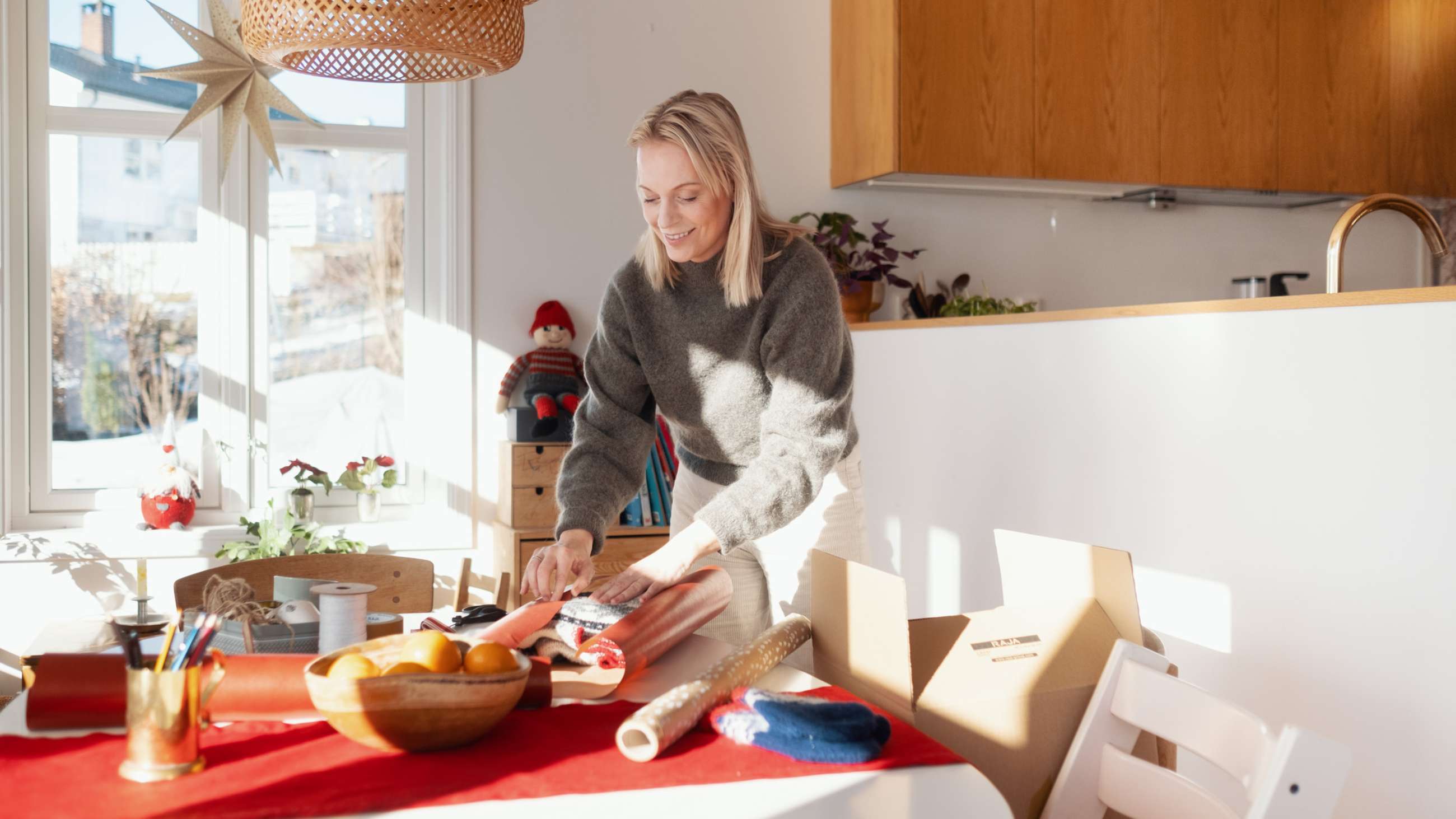 En kvinne lener seg smilende over et bord og pakker en genser i julegave