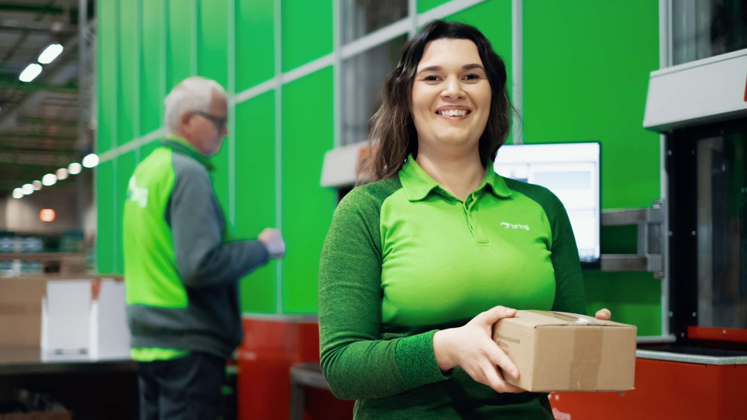 Kvinne kledd i grønne Bring-klær smiler mot kamera mens hun står med pakke i hånden ved en Autostore-stasjon.