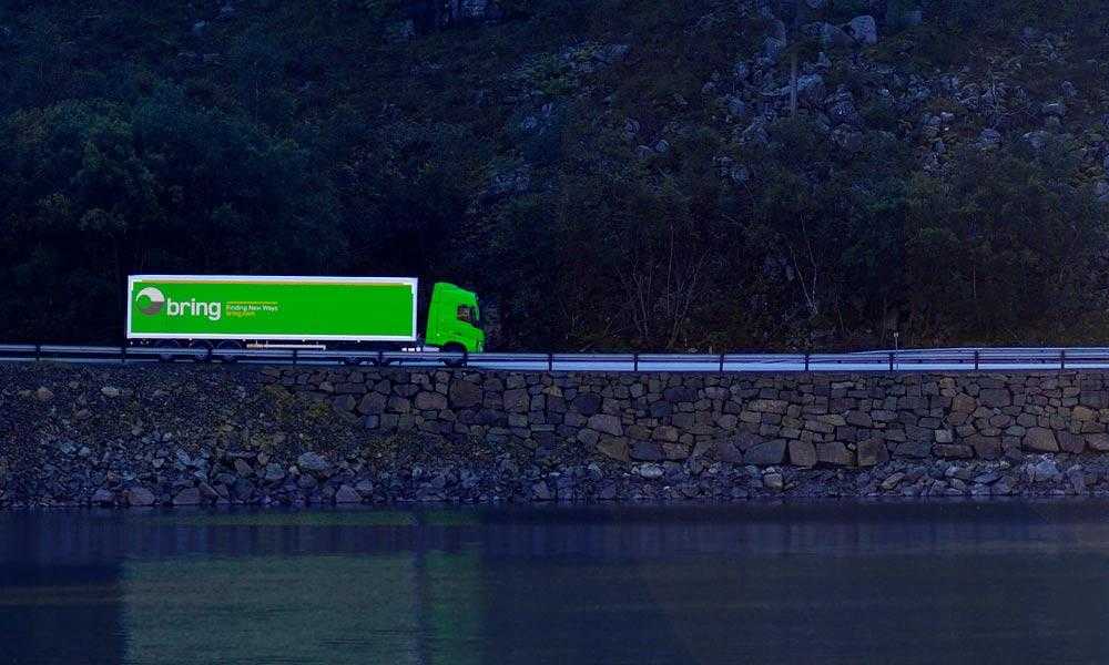 grønn bring lastebil kjører på landsvei langs et vann.