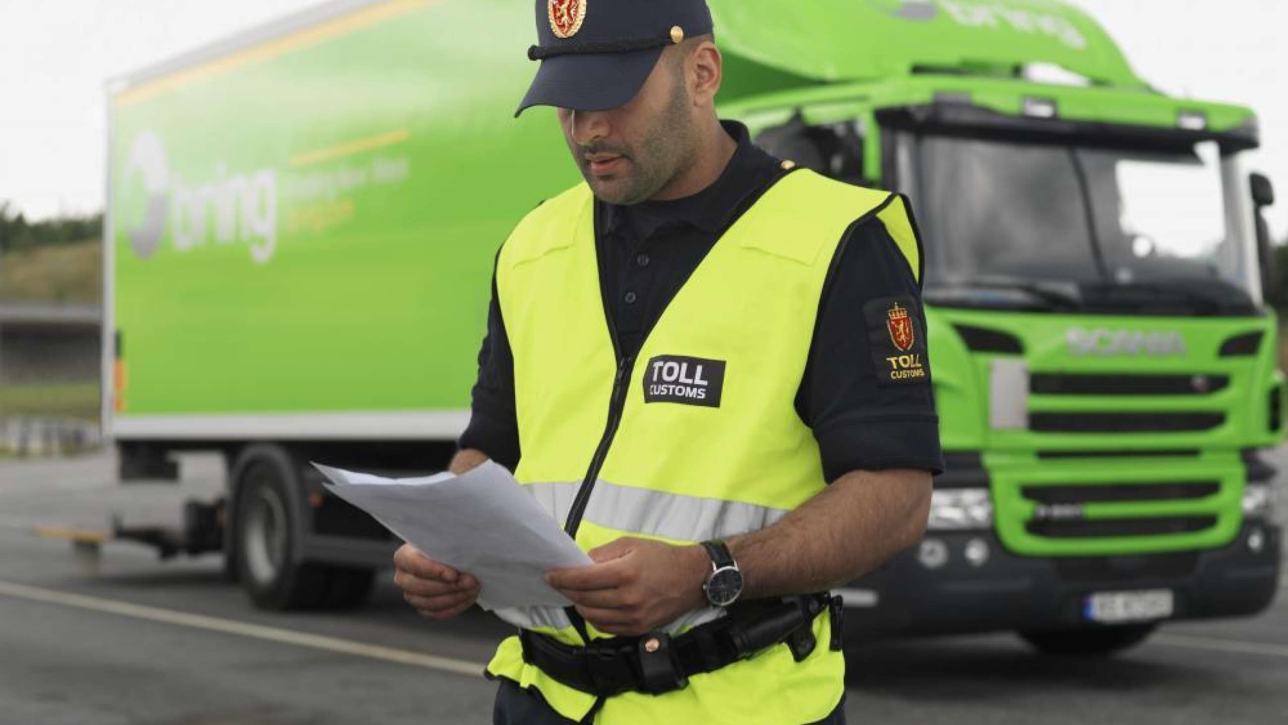 A customs officer checks documents in front of a green truck from Bring.